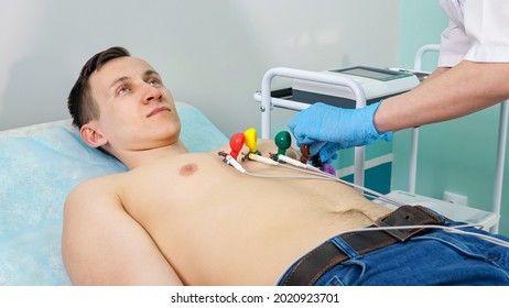 Nurse In Gloves Puts Electrodes On Bare Chest Of Young Man Patient Lying On Couch To Do Electrocardiogram In Light Hospital Office Close View