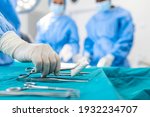 Nurse hand taking surgical instrument for group of surgeons at background operating patient in surgical theatre. Steel medical instruments ready to be used. Surgery and emergency concept