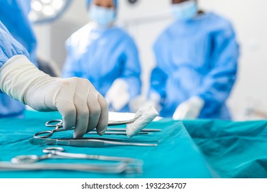 Nurse hand taking surgical instrument for group of surgeons at background operating patient in surgical theatre. Steel medical instruments ready to be used. Surgery and emergency concept