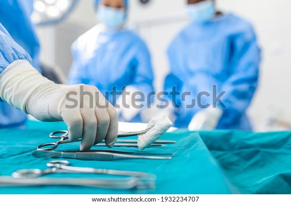 Nurse\
hand taking surgical instrument for group of surgeons at background\
operating patient in surgical theatre. Steel medical instruments\
ready to be used. Surgery and emergency\
concept
