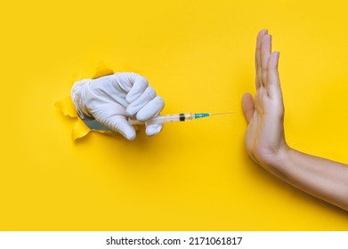 The Nurse Hand In A White Latex Glove Holds A Syringe For Vaccination, But Collides With A Hand In The Form Of A Stop Gesture. Coronavirus Anti-vaccination Concept. Yellow Paper, Torn Hole, Copy Space