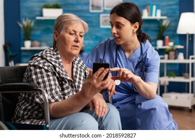 Nurse helping retired senior woman in wheelchair to use smartphone during social service. Elderly person browsing news on digital device in nursing home. Social worker in retirement home - Powered by Shutterstock