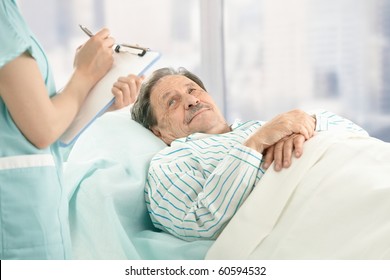 Nurse Holding Clipboard, Taking Notes Of Old Patient Lying In Hospital Bed.?