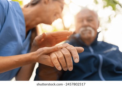 Nurse holding the hand of an elderly man in a wheelchair, both visible in soft focus with a sunlit background, offering comfort and support  - Powered by Shutterstock