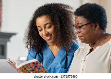 Nurse and lady. Close up of pleasant smiling nurse communicating with lady while reading book for her - Powered by Shutterstock