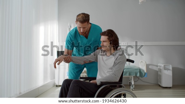 Nurse lifting arm of\
young paralyzed man sitting in wheelchair. Medical worker\
stretching arm and doing exercises with disabled male patient in\
rehabilitation center