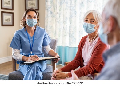 Nurse Listening To Seniors Patients With Face Mask About Covid-19 While Being In A Home Visit. Psychologist During Covid19 Pandemic Wearing Surgical Mask While Visiting Senior Couple At Home.