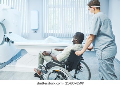 Nurse In Mask Bringing Black Coronavirus Patient In Wheelchair To MRI Procedure In Hospital