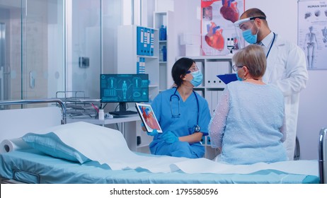 Nurse In Mask With Cardiac Patient At Doctor Appointment Showing A Heart Representation On Digital Tablet In Modern Clinic. Doctor Entering Hospital Room Talking With Old Retired Senior Woman Patient