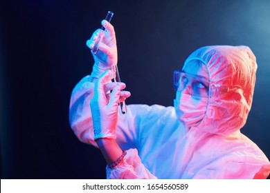 Nurse in mask and white uniform standing in neon lighted room and holding tubes with samples.
