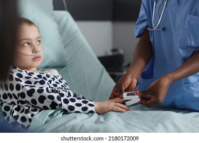 Nurse Measuring Oxygen Levels Of Hospitalized Sick Little Girl Resting On Patient Bed. Ill Kid Sitting In Hospital Pediatric Ward While Medical Staff Monitoring Health Condition Using Oximeter.