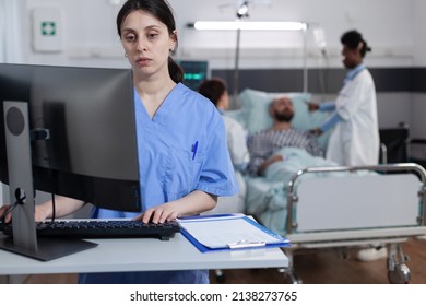 Nurse At Medical Desk Typing Patient Data Into Computer While Team Of Doctors Putting Iv Drip Line On Man With Illness. Health Care Worker With Clipboard Using Pc To Complete Hospital Admission.