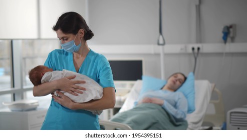 Nurse in medical mask holding newborn baby with mother resting in bed on background. Portrait of female medical worker in protective mask nursing infant kid in hospital ward