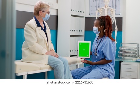 Nurse And Patient With Face Mask Looking At Greenscreen On Digital Tablet, Doing Consultation In Office. Assistant Holding Display With Isolated Chromakey Template And Blank Copyspace.