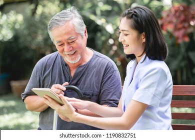 Nurse With Patient Sitting On Bench Together Looking At Tablet. Asian Old Man And Young Woman Sitting Together Talking. Relax Mood.