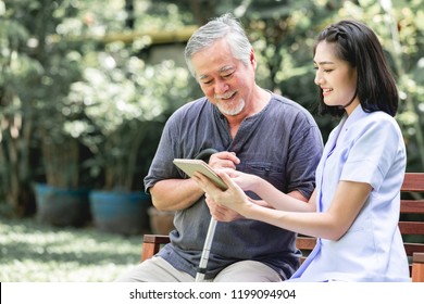 Nurse With Patient Sitting On Bench Together Looking At Tablet. Asian Old Man And Young Woman Sitting Together Talking. Relax Mood.