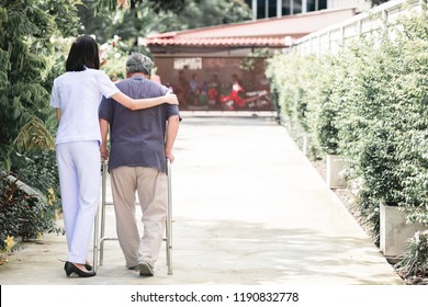 Nurse With Patient Using Walker In Retirement Home. Young Female Nurse Holding Old Man's Shoulder In Outdoor Garden Walking. Senior Care, Care Taker And Senior Retirement Home Service Concept.