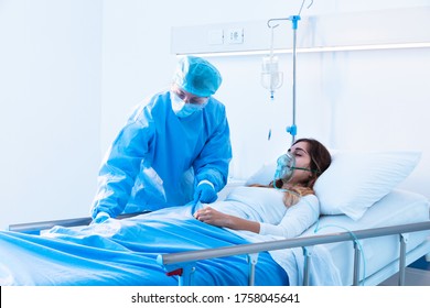 Nurse In PPE Attending To A Covid-19 Patient On The Ward As She Lies In Bed Wearing A Positive Pressure Oxygen Mask To Aid Her Lungs