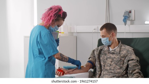 Nurse In Protective Mask Talking To Military Servant Sitting On Chair And Receiving Blood Transfusion At Hospital. Medical Worker With Clipboard Talking To Soldier Blood Donor