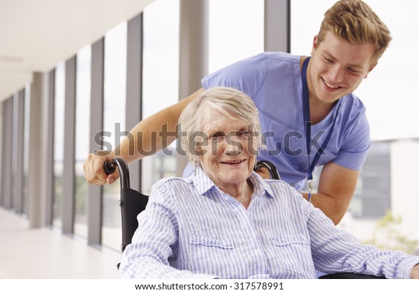Nurse\
Pushing Senior Patient In Wheelchair Along\
Corridor