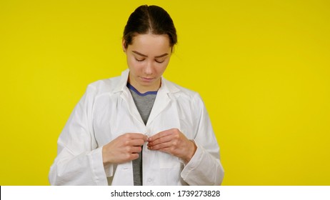 Nurse Puts On A White Coat On A Yellow Background. Young Girl Doctor Getting Ready For Work