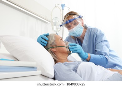 Nurse Puts Oxygen Mask On Elderly Woman Patient Lying In The Hospital Room Bed, Wearing Protective Gloves And Visor Medical Mask, Coronavirus Covid 19 Protection Concept