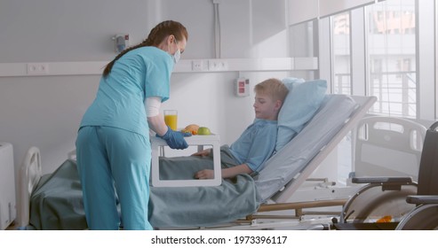 Nurse In Safety Mask And Gloves Serving Child Patient Meal In Hospital Bed. Medical Worker Bringing Lunch Tray For Kid Patient Lying In Hospital Ward 