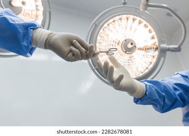 Nurse sending surgical clamp tool for doctor or surgeon inside operating theatre with medical lamp on background.People did surgery.White wall with space.Person in blue gown with yellow light. - Powered by Shutterstock