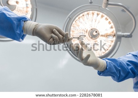 Nurse sending surgical clamp tool for doctor or surgeon inside operating theatre with medical lamp on background.People did surgery.White wall with space.Person in blue gown with yellow light.