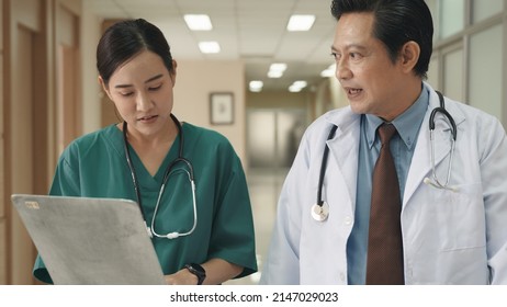 Nurse And Senior Doctor Busy With Clipboard Discussing Along Hospital Corridor, Doctor And Nurse With Medical Report Talking While Walking Down Hallway At The Hospital