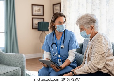 Nurse And Senior Woman Going Through Medical Record On Digital Tablet During Home Visit And Wearing Face Mask. Doctor Wearing Protective Face Mask During Covid Pandemic And Showing Medical Reports.