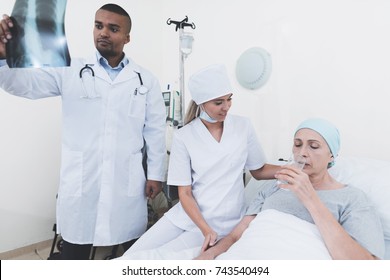 The Nurse Is Sitting Next To The Patient. The Doctor Is Standing Next To A Woman With Cancer, Who Drinks Water. He Looks At Her X-ray. They Are In A Modern Clinic.