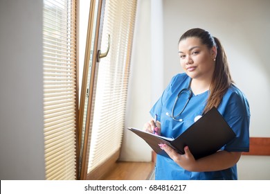 Nurse Standing In Hospital Next To A Window, Holding A Folder And Pen, Looking Into Camera With Straight Face