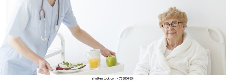 Nurse With A Stethoscope Serving A Diet Meal To Her Elderly Patient Lying In Bed, Wearing A Robe, And Glasses