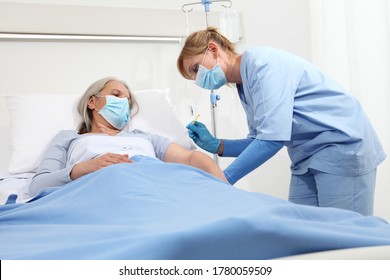 Nurse With The Syringe Injects The Vaccine To The Elderly Woman Patient Lying In The Hospital Room Bed, Wearing Protective Gloves And Medical Surgical Mask, Coronavirus Covid 19 Protection Concept