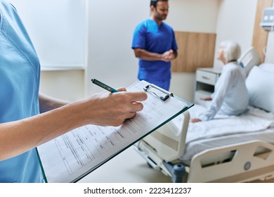 Nurse takes notes in medical chart in hospital room with elderly patient and doctor while bedside consultation. Medical caring about patient - Powered by Shutterstock