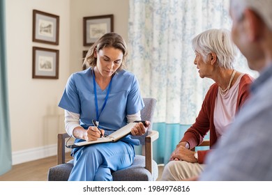 Nurse Talking To Old Patients While Being In A Nursing Home. Young Healthcare Worker Visiting Senior Couple At Care Centre And Writing Information. Nurse In Uniform Noting Symptoms Of Elderly Woman.