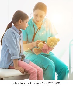 Nurse Teaching Child Patient How To Check Heartbeat With Teddy Bear At Hospital With Yellow Lens Flare In Background