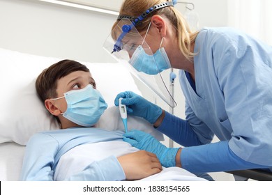 Nurse With Thermometer Measures Fever On Patient Child In Hospital Bed, Wearing Protective Visor And Surgical Mask, Corona Virus Covid 19 Protection Concept, 