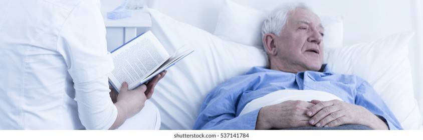 Nurse In Uniform Reading A Book To Elderly Sick Patient Lying In Bed In Hospice And Suffering From Pain