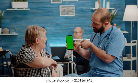 Nurse Vertically Holding Smartphone With Green Screen For Elder Patient With Chronic Disability. Medical Assistant Showing Phone With Chroma Key, Isolated Background And Mockup Template