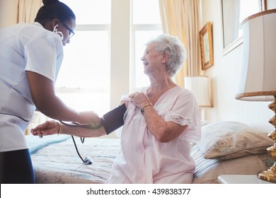 Nurse Visiting Senior Female Patient At Home And Taking Blood Pressure. Old Woman Sitting On Bed.