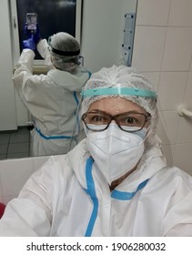 A Nurse Wearing A KN-95 Mask And A Simple Mask, Wearing A Protective White Suit Against Coronavirus, Takes A Selfie In The Break Room, Locker Room Before Work. Real Photo. Real Life.