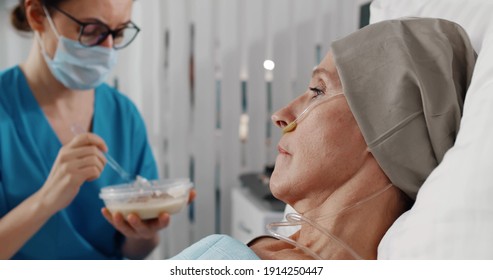 Nurse Wearing Safety Mask Feeding Sick Senior Woman In Hospital. Medical Caregiver In Scrubs Feeding Ill Woman With Cancer Lying In Bed At Hospital Ward