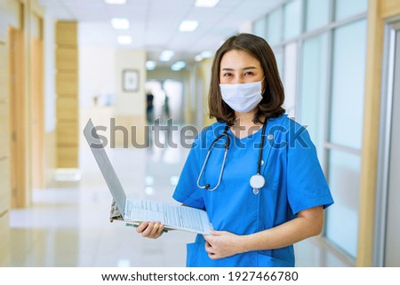 A nurse wearing a white mask looks straight at the camera, standing in front of the corridor. Of hospital clinics

