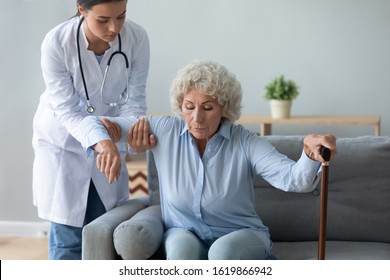 Nurse Wearing White Uniform Coat Helping Older Woman With Walking Cane To Get Up From Couch At Home, Caregiver Supporting Disabled Mature Patient, Rehabilitation And Recovery, Healthcare Concept
