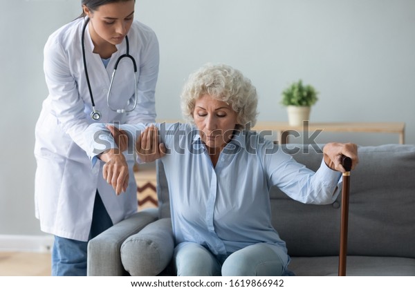 Nurse wearing white uniform coat helping older\
woman with walking cane to get up from couch at home, caregiver\
supporting disabled mature patient, rehabilitation and recovery,\
healthcare concept