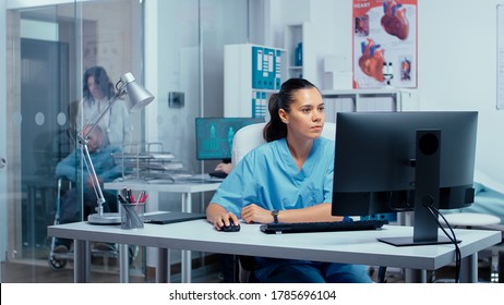 Nurse working on PC in modern private clinic with glass walls where patients are brought in wheelchair from an elevator and other persons are going through hallway. Team of doctors researching - Shutterstock ID 1785696104