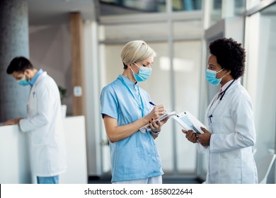 Nurse Writing Notes While Communicating With Black Female Doctor In A Hallway At The Hospital. They Are Wearing Face Masks Due To Coronavirus Pandemic.