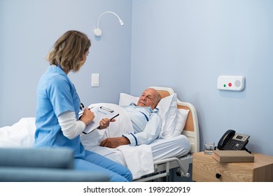 Nurse Writing Recovery Information On Clipboard While Old Patient Lying In Hospital Bed. Young Doctor Writing Medical Notes With Senior Patient In Care Centre. Hospitalized Man Lying On Bed.